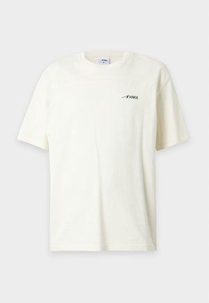 Homme portant un t-shirt blanc uni à manches courtes avec un petit logo noir ARKK sur la poitrine et un pantalon marron, debout devant un fond uni.