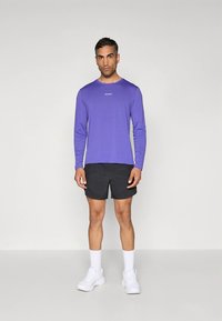 Violetinė ilgomis rankovėmis sportinė palaidinė su logotipu, derinama su juodais šortais ir baltomis sportbačiais. Lygus audinys, įprastas prigludimas, paprastas dizainas.