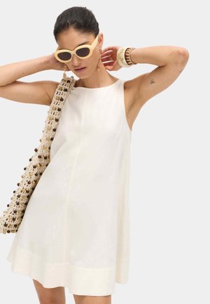 Femme portant une robe blanche sans manches, des lunettes de soleil beige, des boucles d'oreilles en or, des bracelets et portant un sac tricoté avec des perles.