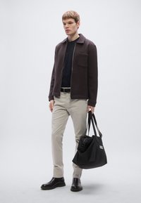 Hnedá bunda na zips, čierne tričko, béžové nohavice, čierne čižmy, drží čiernu tašku s bielym logom. Jednoduchý dizajn, jednoliate farby, moderný strih.