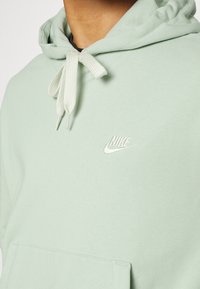 Světle zelená mikina s klokankou, kapucí na šňůrku a vyšívaným bílým logem Nike na hrudi. Měkký, bavlněný materiál.