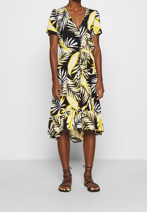 Vrouw draagt een zwarte midi-jurk met geel-witte tropische bladprint, korte mouwen, knopen aan de voorkant en een gerimpelde zoom, gecombineerd met zwarte sandalen.