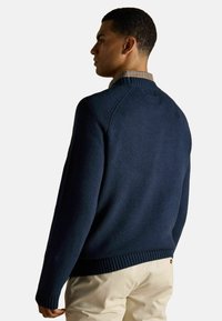 Sinine kootud sviiter ribidega kaela- ja mansettide ning tekstuuriga viimistlusega. Kandmine kontrollitud kaelusega särgi üle, millele on sobitatud heledad püksid.