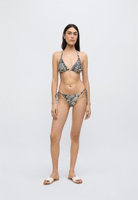 Μπικίνι σε μαύρο και κρεμ με λουλουδάτο σχέδιο· τοπ τρίγωνο με κορδόνια στερέωσης, κάτω μέρος με κορδόνια στα πλαϊνά που ταιριάζει. Το μοντέλο φοράει επίσης λευκές παντόφλες slide.