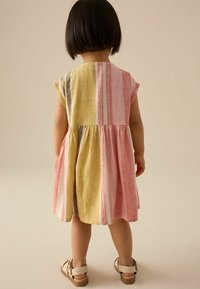 Robe multicolore à manches courtes, comportant des rayures rose, jaune et grise. La jupe est froncée à la taille, au-dessus de sandales.