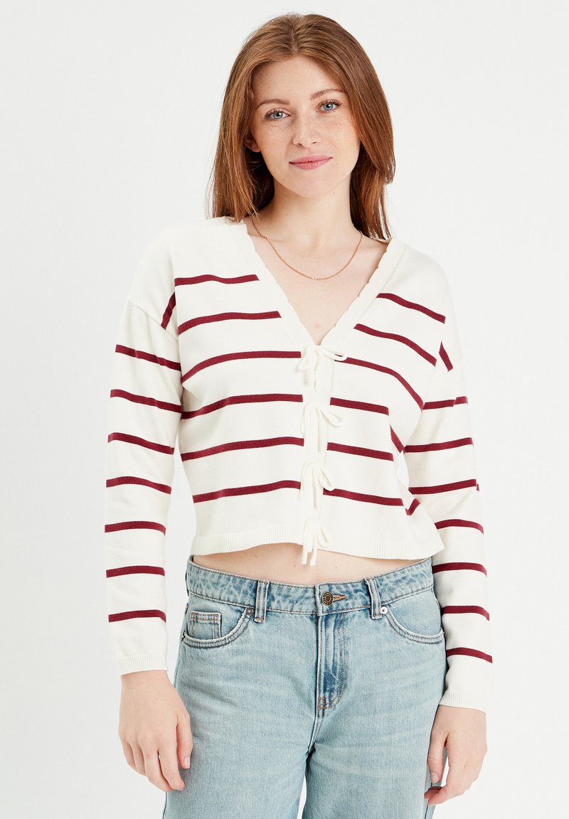 Weißer, verkürzter Pullover mit bordeauxfarbenen Streifen, V-Ausschnitt und gebundenem Detail vorne; die Ärmel sind lang und enganliegend. Kombiniert mit hellblauen Jeans.