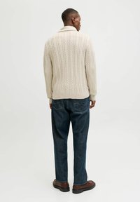 Hellbeige, kabelgestrickter Pullover mit Schalkragen, strukturiertem Muster und geripptem Saum, kombiniert mit lockeren blauen Jeans und dunkelbraunen Schuhen.