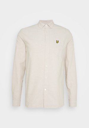 Camisa beige de manga larga con botones y un pequeño logo de mariposa amarilla y negra en el bolsillo izquierdo del pecho.