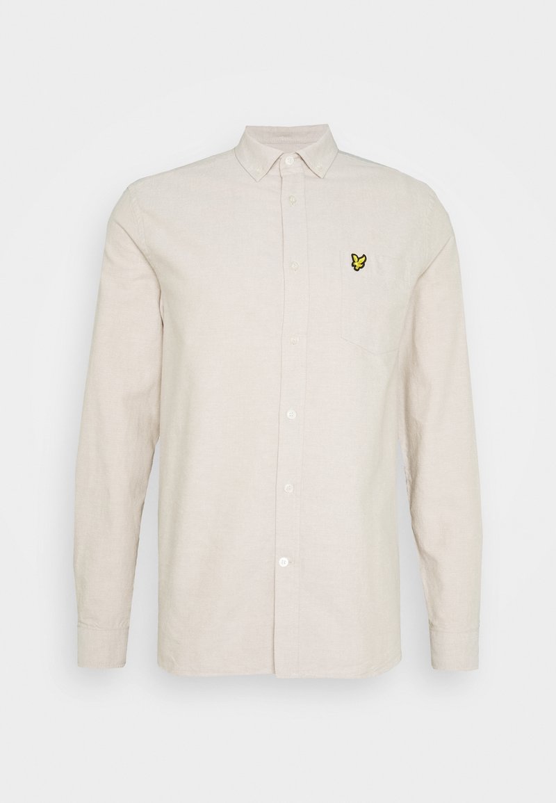 Chemise beige à manches longues boutonnée avec un petit logo papillon jaune et noir sur la poche poitrine gauche.
