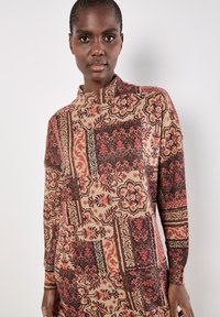 Sweter wykonany na drutach z wzorzystym deseniem w odcieniach czerwieni, brązu i beżu, z wysokim kołnierzem i długimi rękawami, ukazujący skomplikowane faktury.
