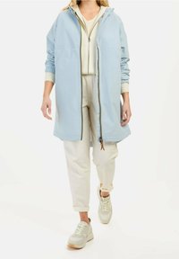 Jasnoniebieska, długa kurtka z kapturem, zapinana z przodu na zamek, z elastycznymi mankietami i dwoma bocznymi kieszeniami. Połączona z kremowymi spodniami i tenisówkami.