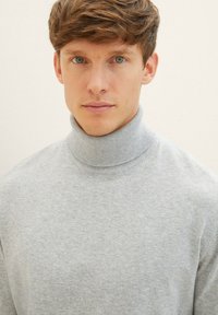 Pull encolure haute gris en tissu doux, avec une texture subtile, porté par un modèle aux cheveux courts.