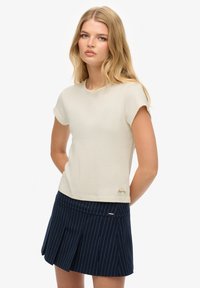 Camiseta de manga corta color crema claro con escote redondeado y un sutil logotipo en el dobladillo, combinada con una falda plisada a rayas marino.