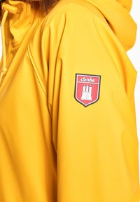 Giacca impermeabile gialla con un patch logato raffigurante un castello. Realizzata in materiale liscio e impermeabile, con cappuccio regolabile e cordini.