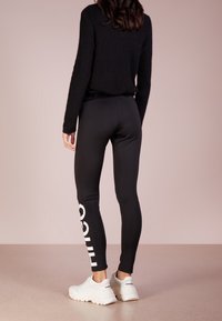 Mustat legginsit, joissa on sileä pinta ja valkoinen logodetail vasemmassa lahkeessa, yhdistettynä pörröiseen mustaan neuleeseen ja valkoisiin tennareihin.