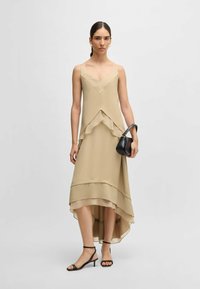 Vaalean beige mekko, jossa on virtaava, kerroksellinen muotoilu, V-pääntie ja epäsymmetrinen helma. Yhdistetty mustiin hihnakenkiin ja pieneen käsilaukkuun.