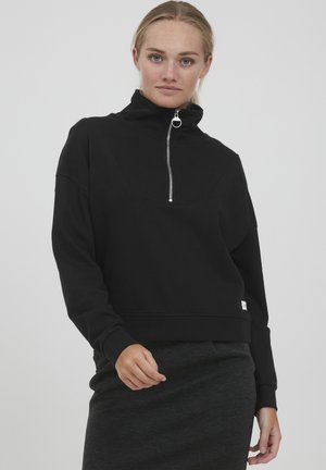 Svart hettejakke med glidelås, høy krage, teksturert stoff og kort design. Sølvglidelås og logo-tag ved kanten gir detaljer.