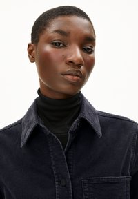 Marineblaues Cord-Jackett mit Kragen, Knopfverschluss und Taschen auf der Vorderseite, über einem schwarzen Rollkragenpullover getragen. Glatte Textur, strukturiertes Design.