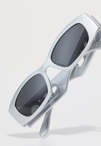 Ochelari de soare cu ramă argintie mată, lentile ovale mari de culoare gri închis și un design elegant, cu accente decupate pe brațele ochelarilor.