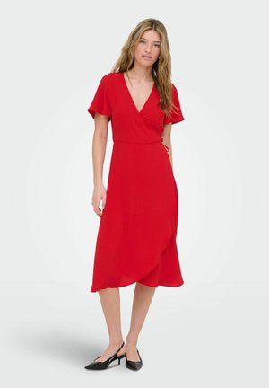 Kvinde står iført en knælængde rød slå-om-kjole med korte ærmer og sorte lavhælede slingback-sko på en ensfarvet baggrund.