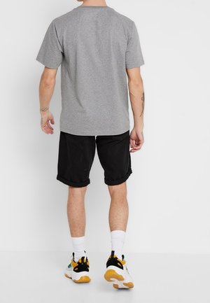 Mann, der ein graues kurzärmeliges T-Shirt, schwarze hochgekrempelte Shorts, weiße Socken und weiße Sneakers mit schwarzen und gelben Akzenten trägt, von hinten gesehen.