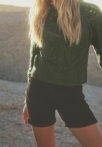 Pull en maille vert foncé avec des motifs de feuilles texturés, associé à un short noir retroussé. Le pull a une coupe décontractée et un ourlet côtelé.