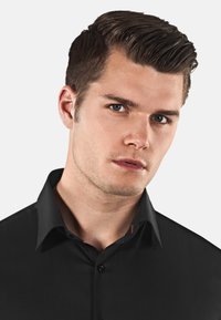 Zwarte button-up shirt met een puntkraag, voorzien van een rode accentlabel. Gladde textuur en een fitted design, gericht op de halslijn en schouders.