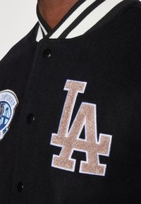 Zwarte wollen varsityjack met een witte en zwarte gestreepte kraag, knoopsluiting en een geborduurd "LA"-logo in bruin en blauw.