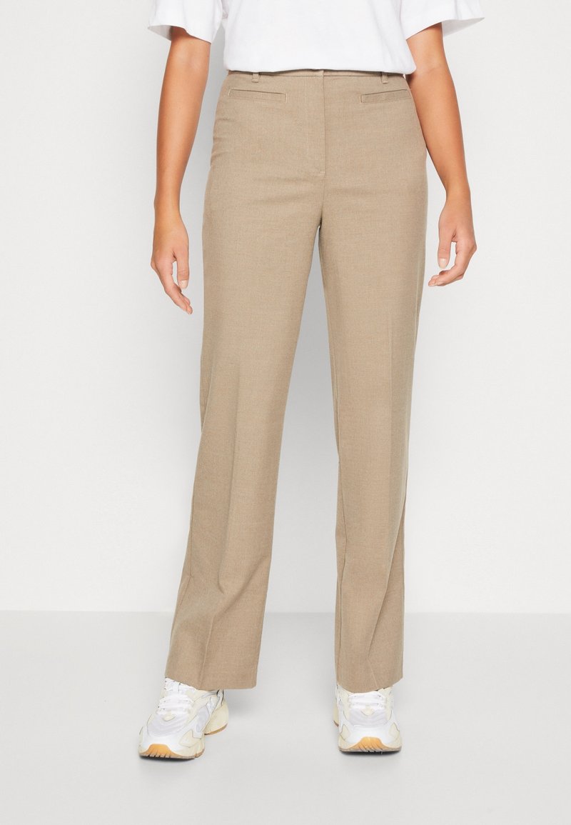 Personne portant un pantalon beige droit et des baskets blanches, debout devant un fond blanc uni.
