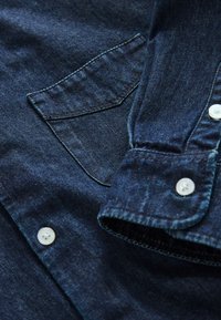 Gros plan d'une chemise en denim bleu foncé montrant une poche avant, un poignet avec bouton et des boutons blancs sur la texture du tissu.