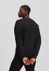 Sweatshirt negru cu decolteu rotund, mâneci lungi și manșete ribbate. Textură moale, design simplu. Modelul prezintă vederea din spate.