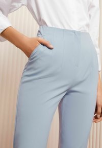 Világoskék, testre szabott nadrág sima textúrával, slim fit szabással, oldalsó zsebekkel és diszkrét redőzéssel az elején. Fehér inggel párosítva.