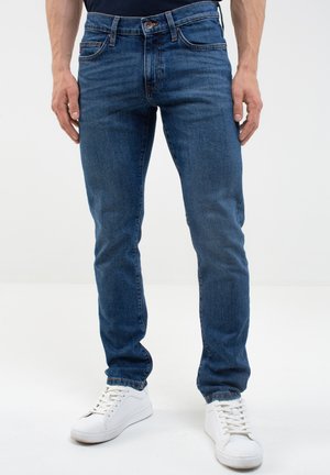 Jeans de mezclilla azul con un corte slim, que presentan un clásico diseño de cinco bolsillos, sutil deslavado y cierre de botón y cremallera. Combinados con zapatillas blancas.