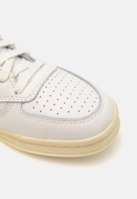 Weiße Ledersneaker mit perforiertem Zehenteil, cremefarbener Gummisohle, hellblauen Akzenten und flachen Schnürsenkeln. Glatte Textur und abgerundete Form.