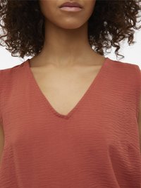 Donna con capelli ricci che indossa una maglietta smanicata color terracotta con scollo a V, che mostra il collo e la parte superiore del petto.