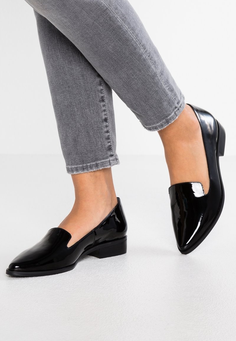 Jambes portant un jean gris coupé et des mocassins noirs en cuir brillant à enfiler avec un petit talon sur un sol blanc.
