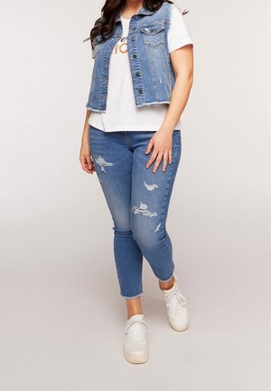 Kvinde stående iført hvide sneakers, revnede blå jeans, hvid T-shirt og ærmeløs denimvest mod ensfarvet baggrund.