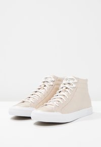 Zapatillas altas de cuero beige claro con suelas de goma blancas, que presentan cordones blancos y detalles de costura sutiles en la parte superior.