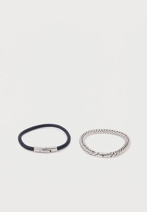 Deux bracelets sur un fond blanc : l'un est en cuir noir tressé avec un fermoir en argent, l'autre est un bracelet à maillons en argent avec un fermoir.