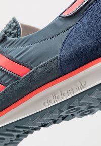 Buty sportowe Adidas z mieszanką szarego materiału tekstylnego i zamszu, czerwonymi paskami oraz białą podeszwą z wzorami bieżnika; teksturowane akcenty na cholewce.