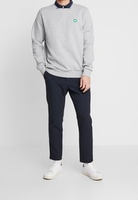 Felpa grigia con polsini a costine, dotata di un piccolo logo verde. Indossata sopra un polo blu navy con colletto bianco, abbinata a pantaloni blu navy e sneakers bianche.