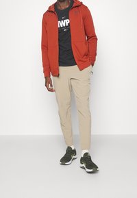 Red zip-up hoodie over a black t-shirt, beige joggers, and green sneakers with white soles. Smooth fabric and relaxed fit.
