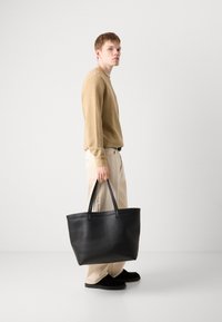 Mala tote de couro preto com textura suave, laterais altas e duas alças robustas. Usada por uma pessoa com um suéter bege e calças claras.