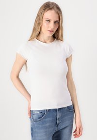 Λευκό κοντομάνικο μπλουζάκι με στρογγυλή λαιμόκοψη και ελαφρώς φθαρμένες άκρες, συνδυασμένο με μπλε τζιν παντελόνι. Απλός, στενός σχεδιασμός.