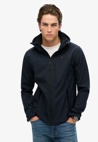 Giacca softshell blu navy con chiusura a zip, multiple tasche e cappuccio. Caratterizzata da una vestibilità slim e tessuto strutturato. Abbinata a jeans in denim.