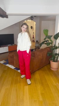 Balta ar rāvējslēdzēju stiprināma flīsa jaka, sarkanas sporta bikses un bēšas čības. Stāv dienas istabā ar koka grīdu, brūnu dīvānu un istabas augiem.