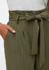 Olijfgroene, getextureerde stoffen broek met een brede tailleband, voorplooien en een strikaccent. Zak zichtbaar aan de zijkant.