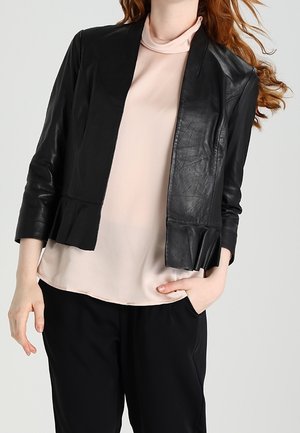 Kobieta nosząca czarną skórzaną kurtkę z rękawami do łokcia na jasnoróżowej bluzce i czarnych spodniach, stojąca z jedną ręką w kieszeni.