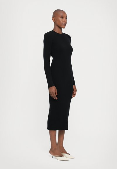 Rochie lungă din tricot cernit, cu mâneci lungi și o siluetă ajustată, care ajunge până la jumătatea gambei. Este purtată cu mocasini de culoare crem, cu vârful ascuțit.