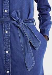Denimkjole i mørk blå med knappelukking foran, lange ermer og en knyttet midjebelt. Har synlige sømmer og lommer på sidene.